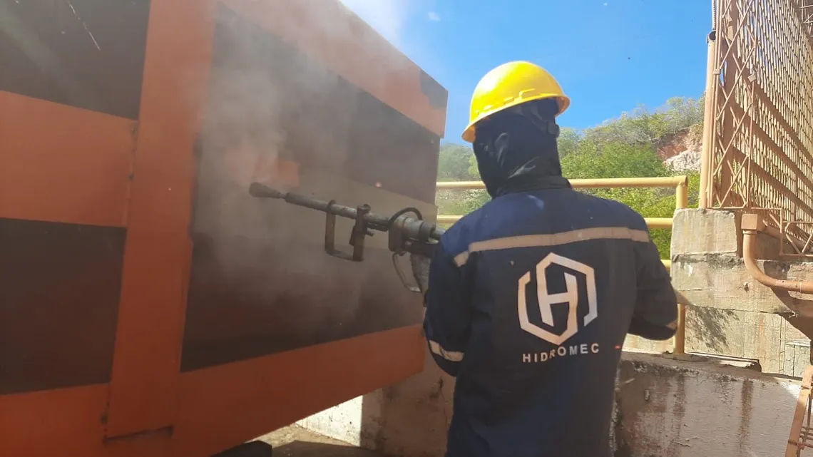 Operador de uniforme azul com o logotipo HIDROMEC e capacete amarelo aplicando lança de hidrojateamento sobre uma comporta metálica alaranjada.
