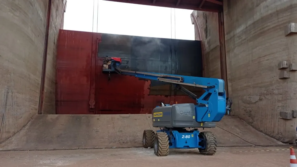 Plataforma elevatória azul posicionada dentro do vão do vertedouro, com operador aplicando hidrojateamento sobre a face de uma comporta vermelha.