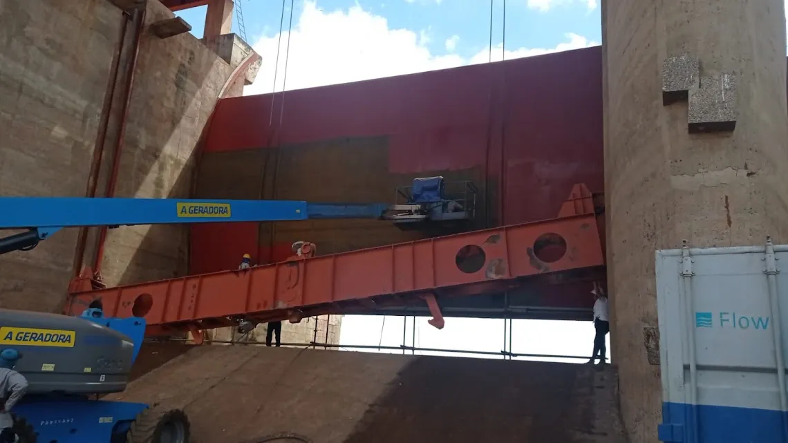 Vão de vertedouro com viga metálica alaranjada suspensa, plataforma elevatória e contêiner de equipamento Flow ao lado direito.