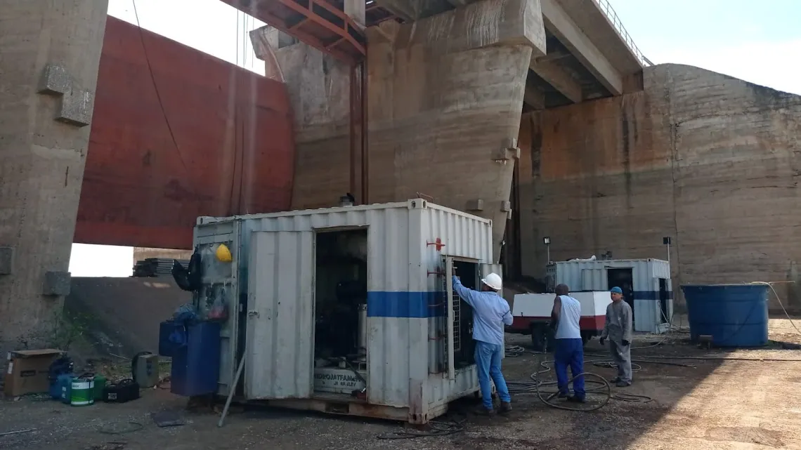 Equipe posicionando contêiner branco e azul da unidade de hidrojateamento ao pé da estrutura de concreto do vertedouro.