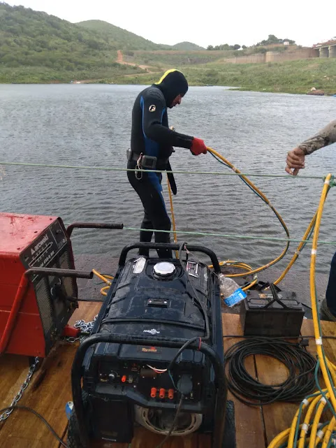Mergulhador em roupa de neoprene sobre balsa de madeira, ajustando mangueiras junto a um gerador e compressor, com o reservatório ao fundo.