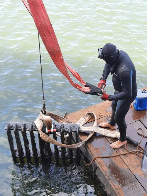 Mergulhador em roupa de neoprene sobre balsa, prendendo cintas de içamento a uma grade metálica coberta de limo retirada da água.