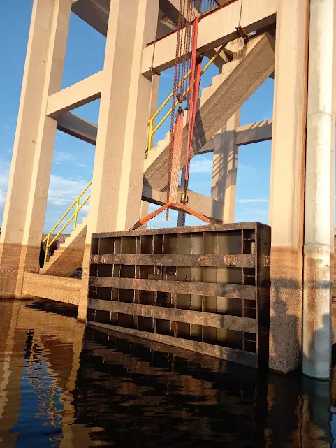 Comporta stop-log suspensa por talha de corrente entre os pilares de concreto da tomada d'água, sobre a água do reservatório.
