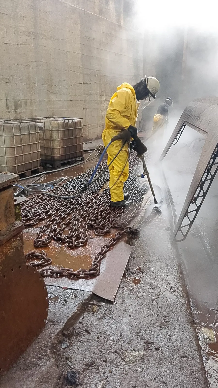 Operador com vestimenta impermeável amarela, capacete e protetor facial aplicando lança de hidrojateamento, com correntes de aço sobre o piso e névoa de água ao lado.