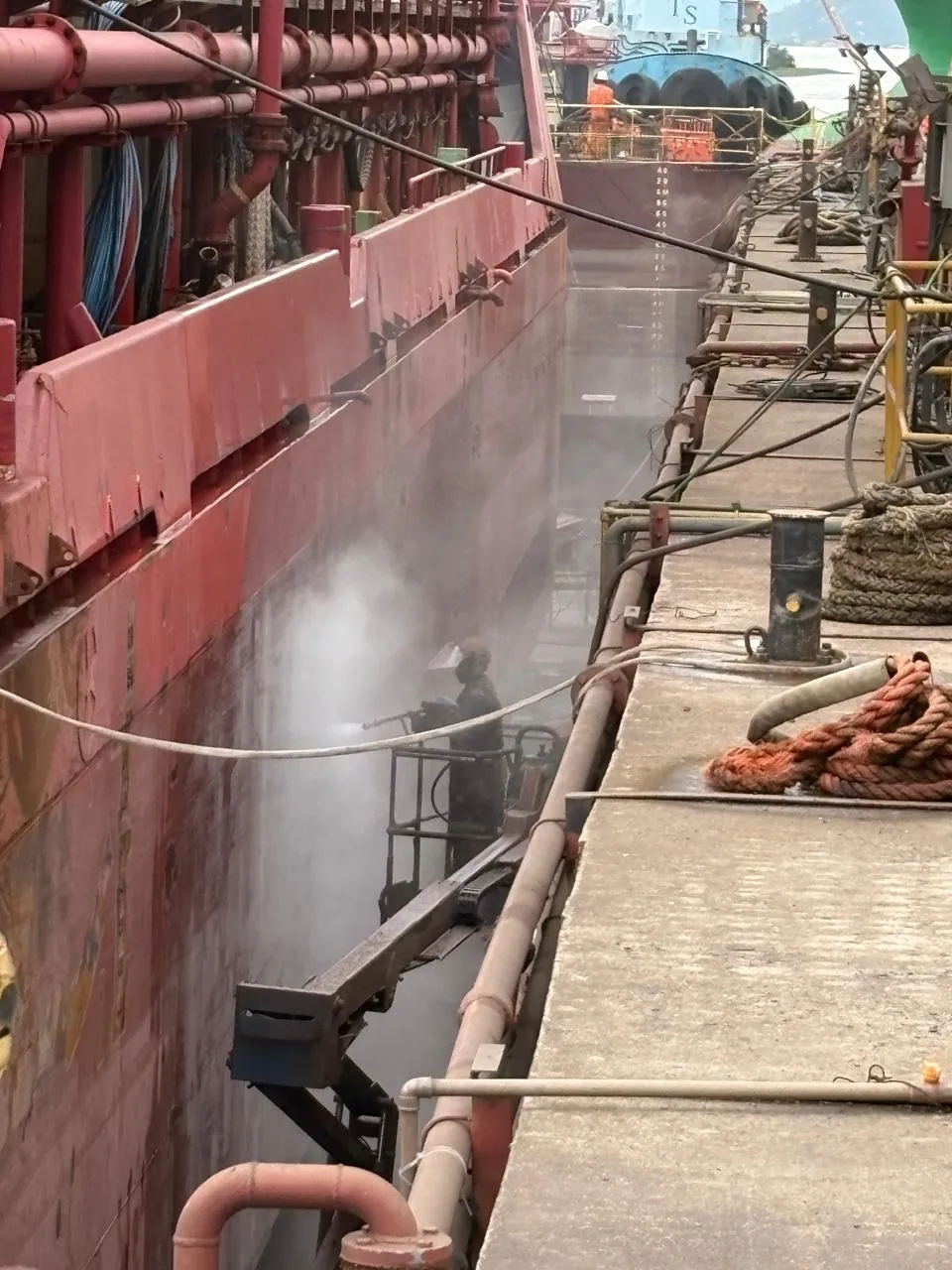 Operador em plataforma suspensa aplicando hidrojateamento ao lado do casco vermelho de uma embarcação atracada, com cais, cabeço de amarração e cabos à direita.