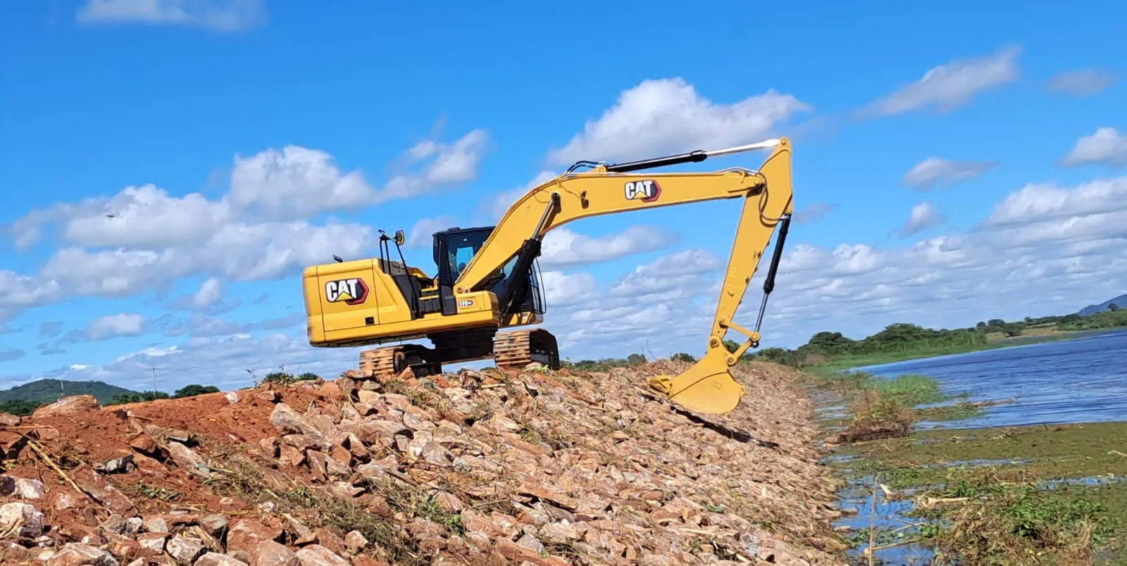 Escavadeira Caterpillar posicionada sobre o talude lançando pedra de enrocamento, com o reservatório de Angicos à direita.