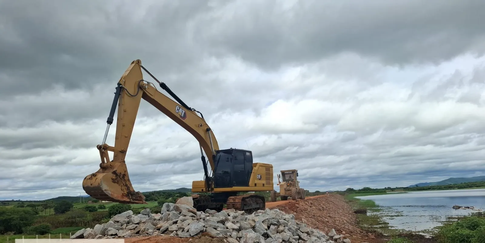 Escavadeira Caterpillar sobre a crista da barragem ao lado de pilha de pedra de enrocamento, com motoniveladora ao fundo e o reservatório à direita.