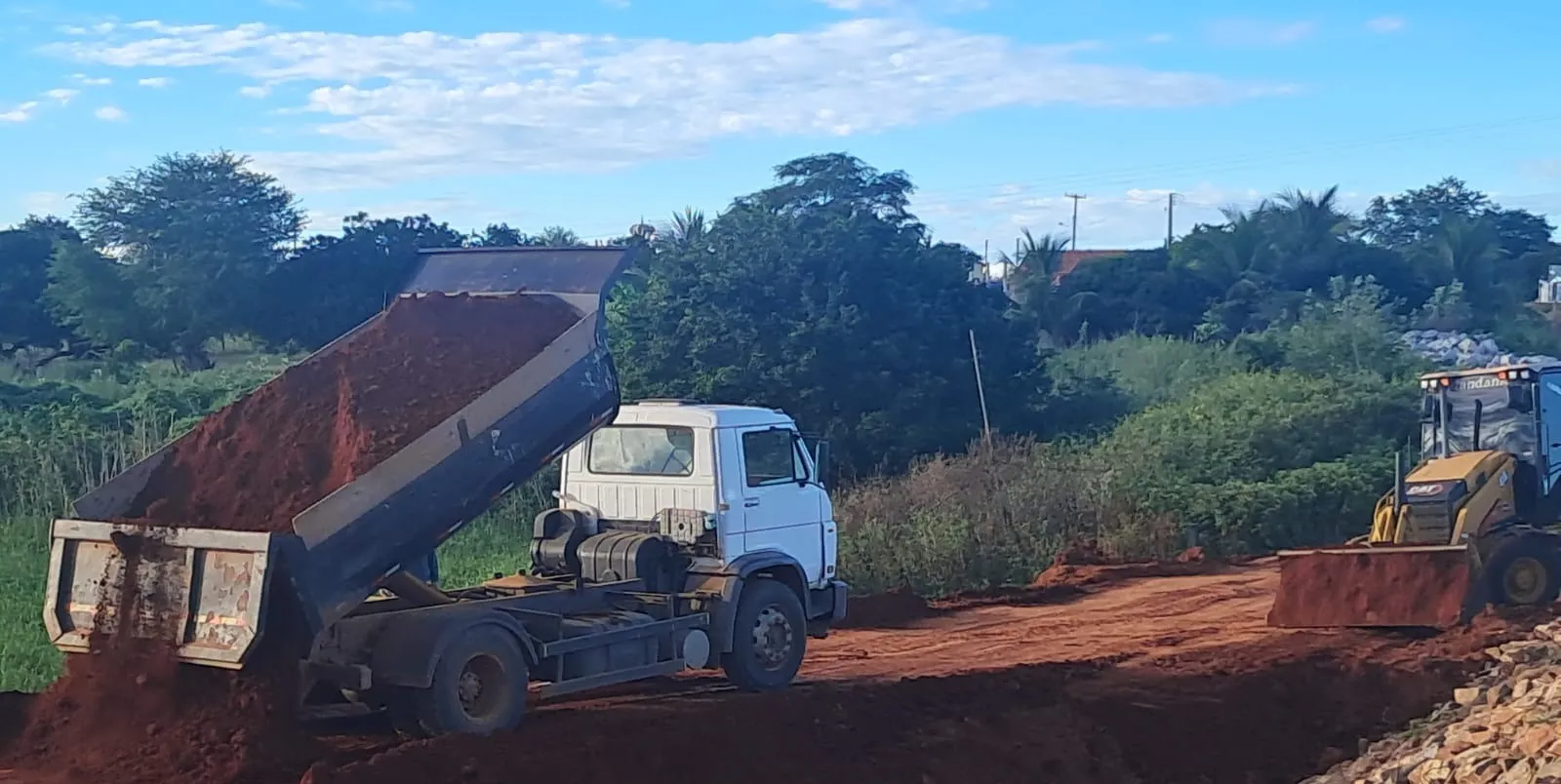 Caminhão basculante descarregando solo vermelho para a formação da ensecadeira, com pá-carregadeira Caterpillar espalhando o material.