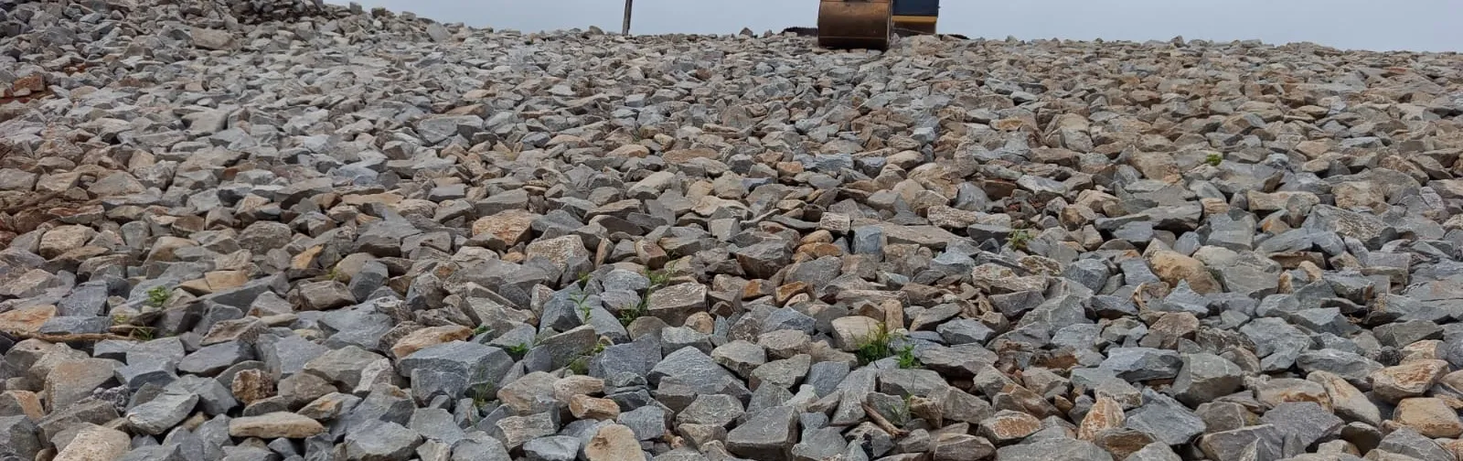 Talude coberto por enrocamento de pedra em toda a extensão, com escavadeira trabalhando na crista ao fundo.