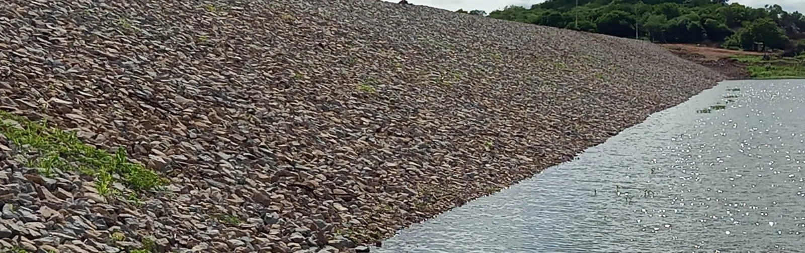 Talude de montante revestido com enrocamento de pedra descendo até a água do reservatório de Lagoa do Arroz.