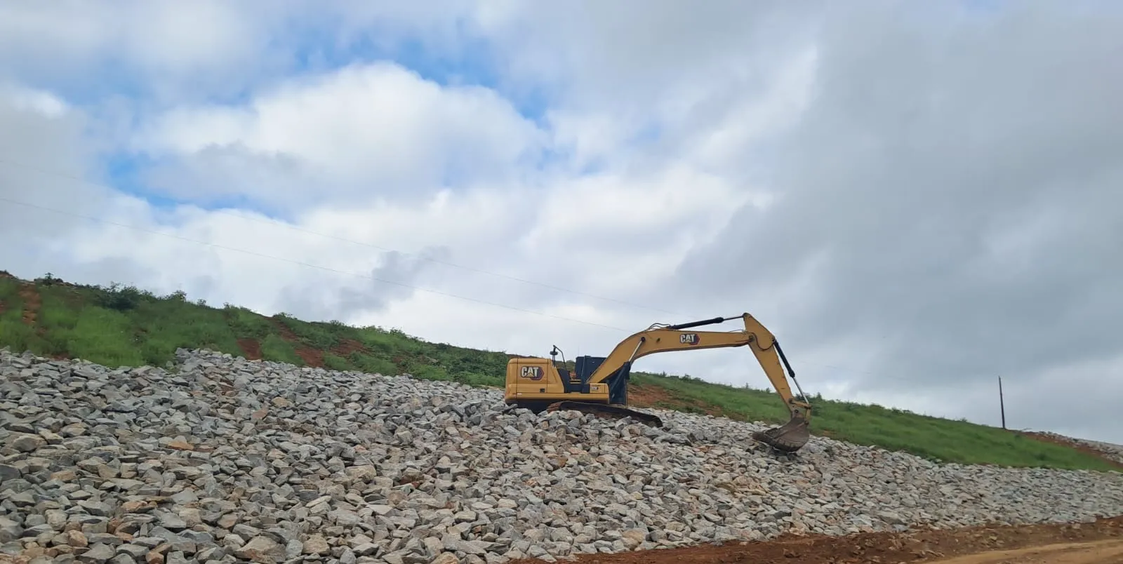 Escavadeira Caterpillar posicionada sobre a pedra recém-lançada no talude de jusante, com caminho de serviço em primeiro plano.