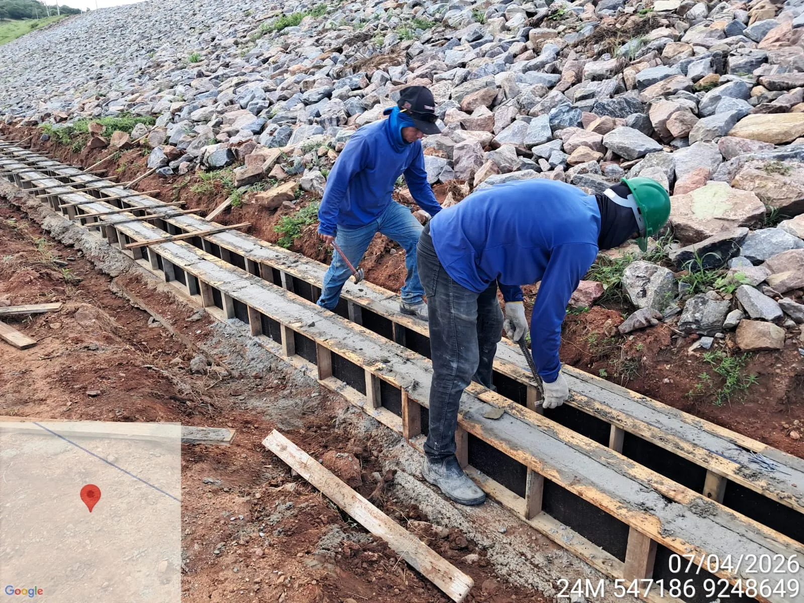 Dois trabalhadores concretando calhas de drenagem em formas de madeira ao pé do talude de enrocamento.