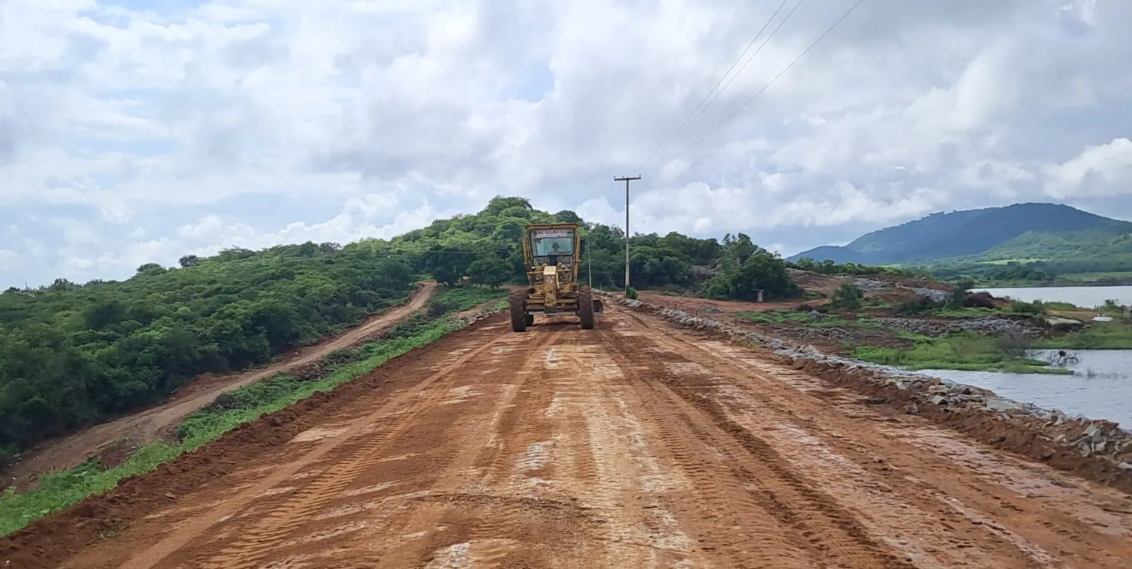Motoniveladora nivelando o coroamento de solo da barragem, com enrocamento à direita e o reservatório ao fundo.