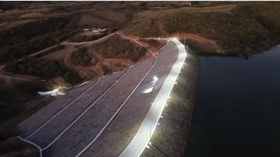 Vista aérea noturna da Barragem dos Prazeres com o coroamento iluminado por postes ao longo de toda a crista e o talude de enrocamento à esquerda.