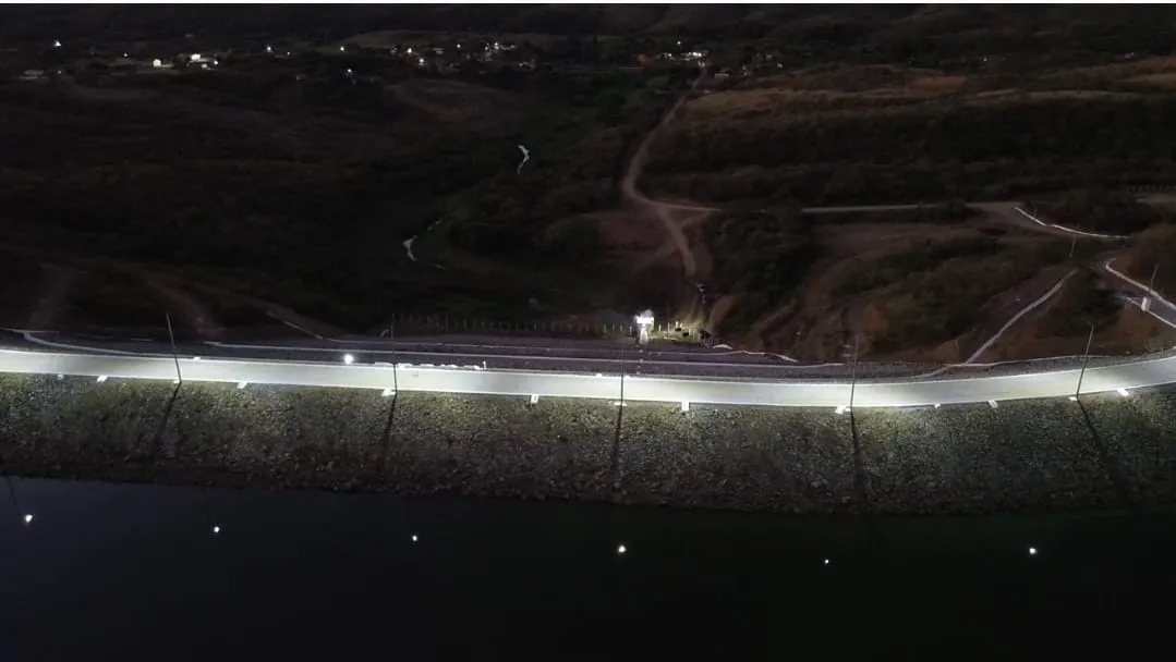 Vista aérea noturna do coroamento da Barragem dos Prazeres, com a faixa de iluminação percorrendo a crista e o reservatório escuro à frente.