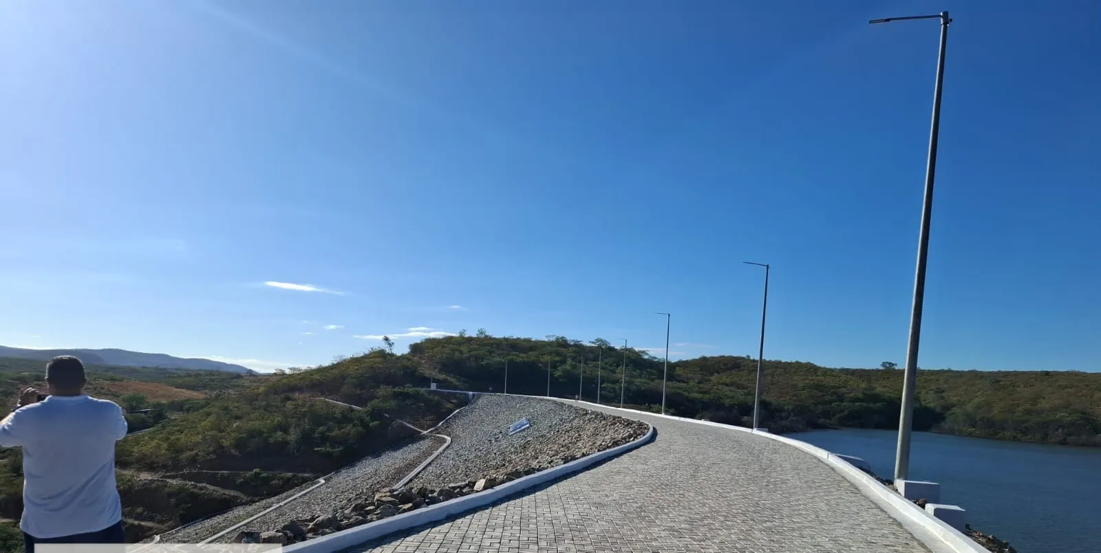 Coroamento da barragem revestido com piso intertravado, meio-fio pintado de branco e postes de iluminação, com o reservatório à direita.