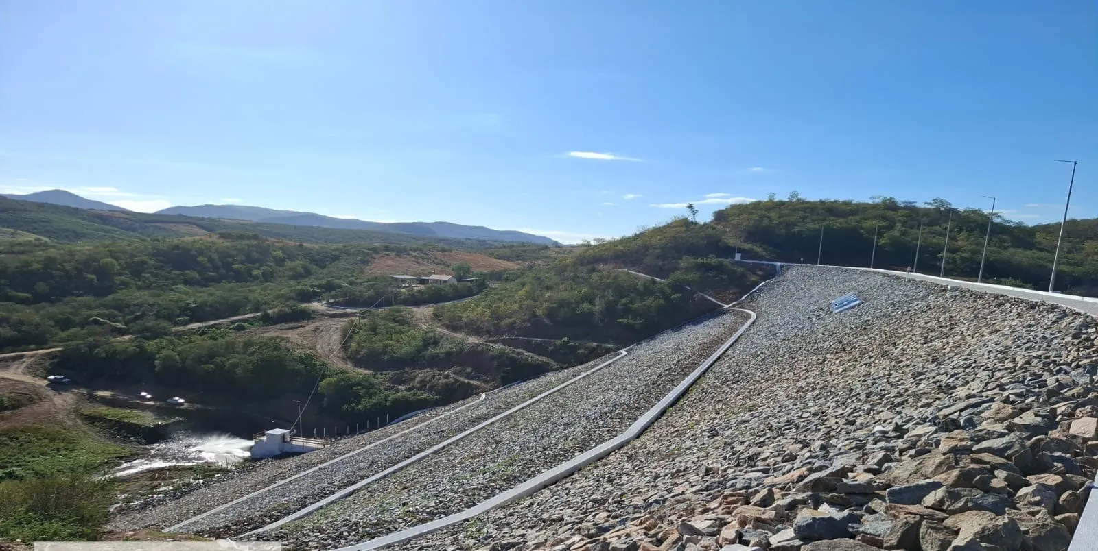 Talude de jusante da Barragem dos Prazeres com enrocamento de pedra e três calhas de drenagem em concreto descendo a encosta.
