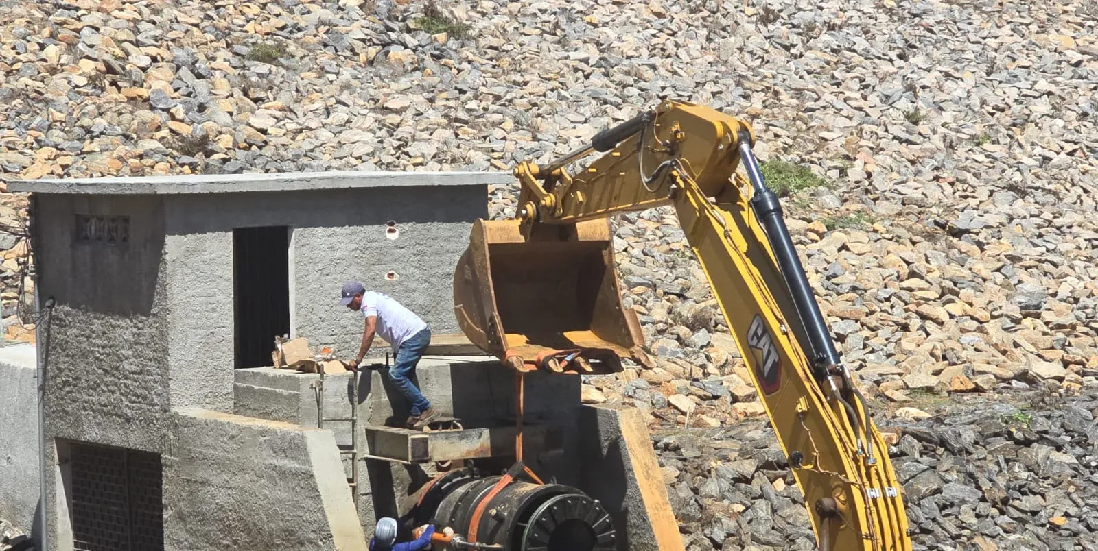 Escavadeira Caterpillar posicionando a válvula dispersora sobre a estrutura de concreto, com dois trabalhadores guiando a peça.
