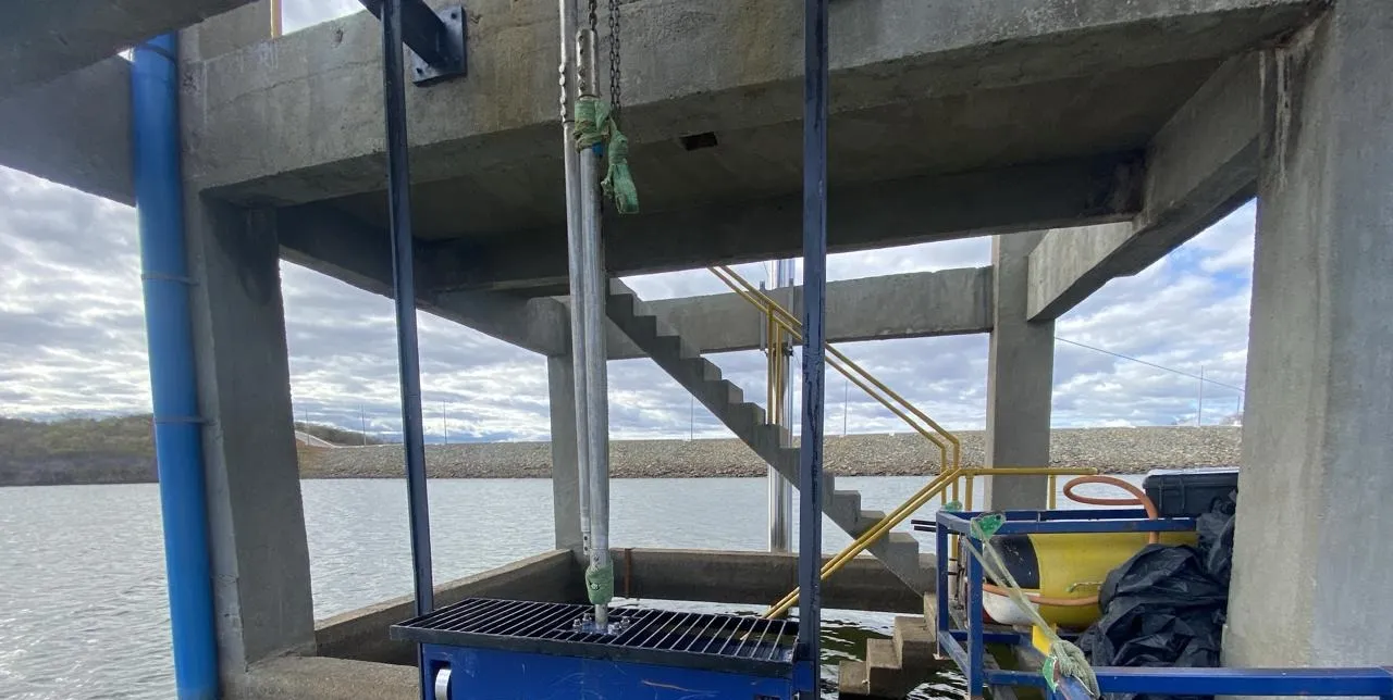 Comporta vagão pintada de azul sendo posicionada por talha sob a estrutura de concreto da tomada d'água, sobre o reservatório.