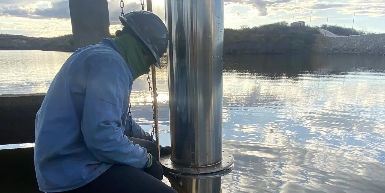Trabalhador de capacete cinza fixando o flange de um tubo de aeração em aço inoxidável junto à torre da tomada d'água, ao pôr do sol.