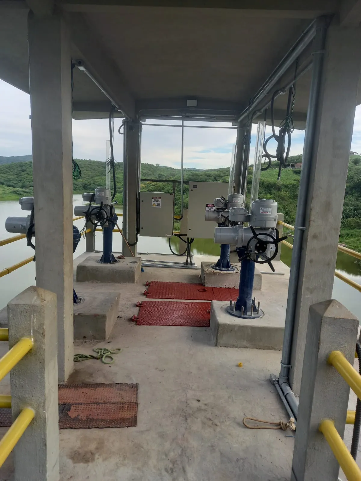 Plataforma coberta da torre de manobra com quatro atuadores elétricos instalados sobre hastes azuis de acionamento de comportas, com o reservatório ao fundo.